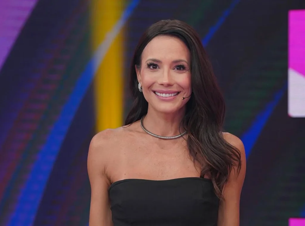 Juliana Knust sorridente, com cabelos castanhos longos e ondulados, usando um vestido preto tomara que caia e um colar rígido prateado em um cenário colorido de estúdio de televisão.