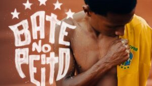 Homem negro de perfil, com a pele suada, segurando a gola de uma camisa amarela da Seleção Brasileira de Futebol. Ao lado esquerdo, há a inscrição estilizada em letras brancas 'Bate No Peito' cercada por estrelas, sobreposta a um fundo em tom terroso.