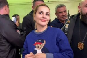 Advogada e influenciadora digital Deolane Bezerra sorri levemente, vestindo um moletom de tricô azul escuro com a estampa do urso Ralph Lauren Polo Bear. Ela está em um ambiente movimentado com outras pessoas ao fundo, incluindo homens que parecem fazer sua segurança.