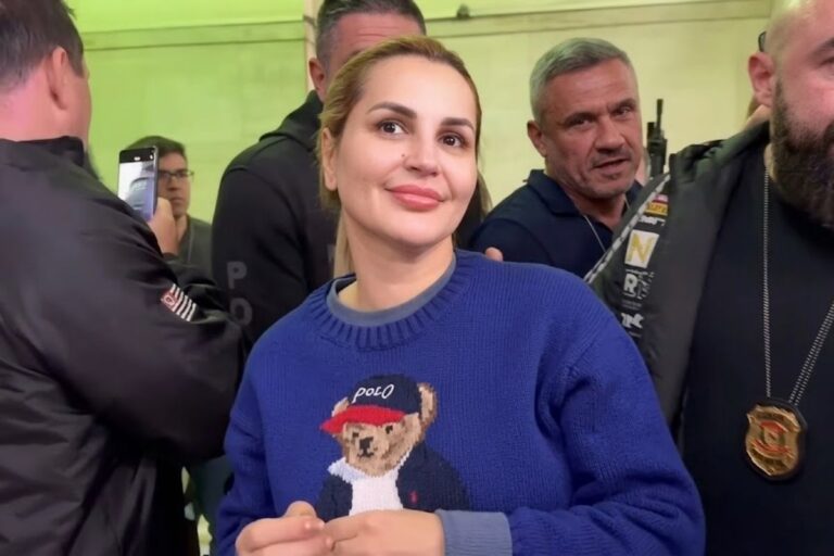 Advogada e influenciadora digital Deolane Bezerra sorri levemente, vestindo um moletom de tricô azul escuro com a estampa do urso Ralph Lauren Polo Bear. Ela está em um ambiente movimentado com outras pessoas ao fundo, incluindo homens que parecem fazer sua segurança.