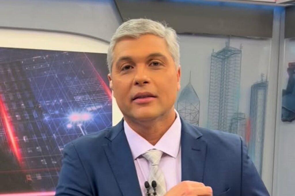 O jornalista e apresentador Joel Datena em primeiro plano, olhando para a frente. Ele tem cabelos grisalhos, veste um paletó azul-marinho sobre camisa social clara e gravata texturizada em tons de cinza. Ao fundo, aparece o cenário de um estúdio de telejornalismo com painéis digitais ilustrando prédios e gráficos.