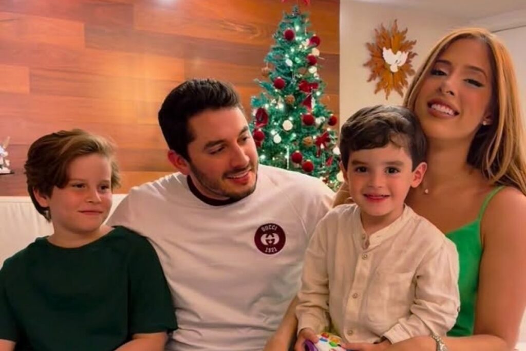 O cantor Jonas Esticado, com camisa branca de manga longa e emblema, está sentado em um sofá. À sua esquerda está seu filho Mikael, com camisa verde escura. À sua direita, sua esposa, Bruna Hazin, com blusa verde alça fina, segura no colo o filho do casal, que usa camisa de botão bege. Atrás, uma árvore de Natal decorada e parede de madeira.