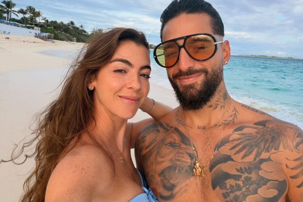 O cantor Maluma, sem camisa e exibindo tatuagens no peito, sorri ao lado de sua namorada, Susana Gómez, em uma praia com areia clara e mar azul ao fundo. Maluma usa óculos de sol grandes com lentes laranjas.