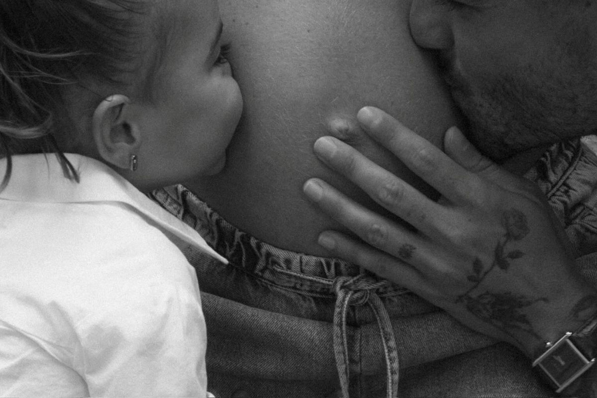 Close em preto e branco de Maluma e sua filha primogênita, Paris, beijando a barriga de gravidez da namorada do cantor, Susana Gómez. A mão tatuada de Maluma repousa suavemente sobre a barriga.
