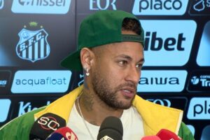 O jogador de futebol Neymar Jr de perfil três quartos, homem pardo, com barba cerrada, usando boné verde virado para trás, brinco de brilhante com pingente de coração e jaqueta verde e amarela sobre camiseta branca, falando ao microfone em frente a um painel com o escudo do Santos Futebol Clube e patrocinadores.