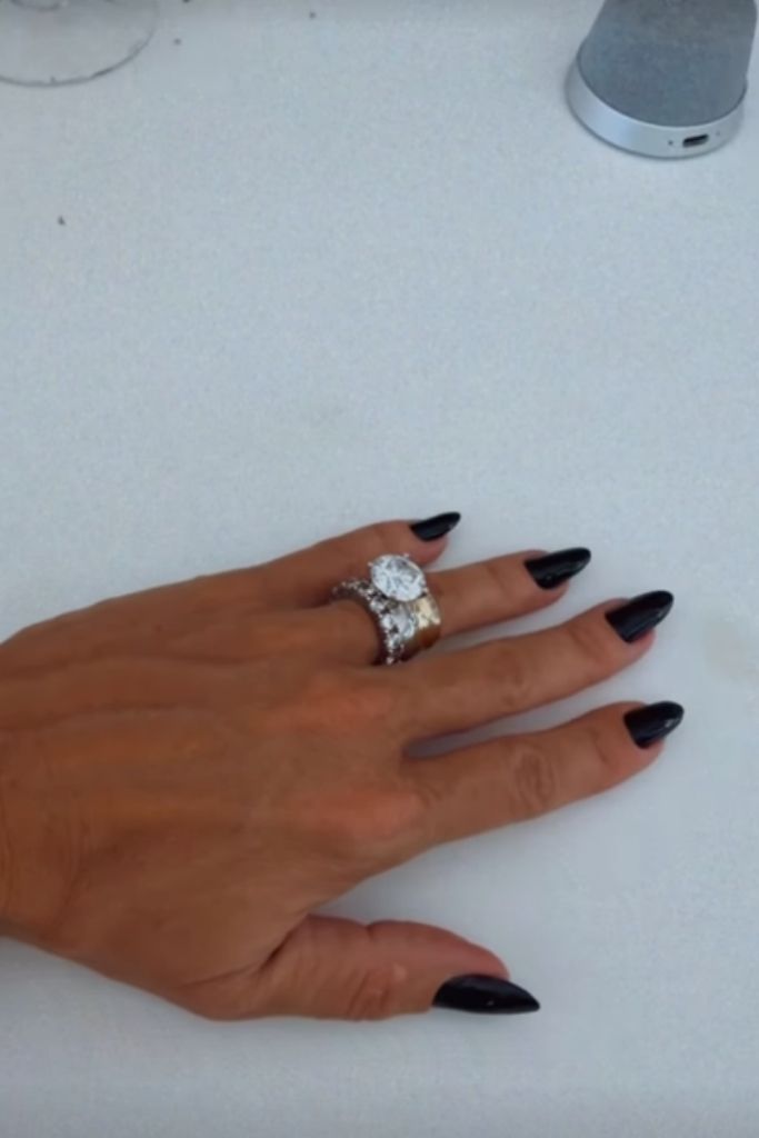 Mão de Poliana Rocha com unhas pintadas de preto, exibindo um anel de luxo com um grande brilhante central e aparadores cravejados de diamantes sobre uma superfície branca.