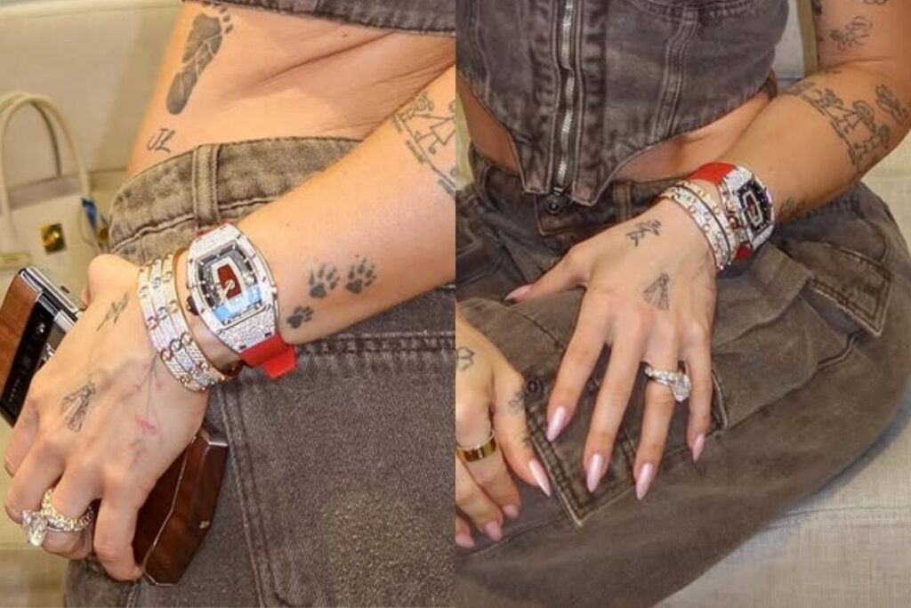Close focado no pulso e nas mãos da influenciadora Virgínia Fonseca, destacando um relógio de luxo cravejado de diamantes com pulseira vermelha e formato tonneau, acompanhado de braceletes de ouro também com brilhantes. Em segundo plano, aparecem suas tatuagens nos braços e mãos, além de um conjunto de anéis e sua roupa em tom marrom texturizado.
