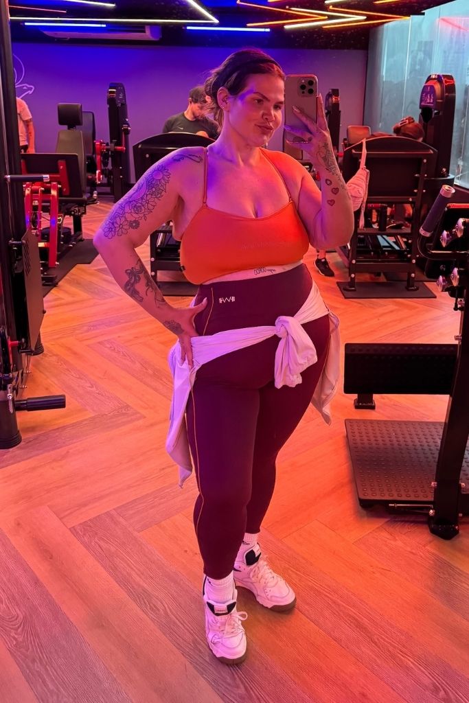 Influenciadora e dançarina Thais Carla em pé, tirando uma foto no espelho de uma academia. Ela veste um top esportivo laranja, calça legging roxa de cintura alta e tênis brancos, com um casaco claro amarrado na cintura. Ela exibe suas tatuagens no braço e posa com a mão esquerda na cintura, segurando o celular com a mão direita. O ambiente ao fundo possui iluminação em tons de roxo e rosa, com aparelhos de musculação e piso amadeirado.