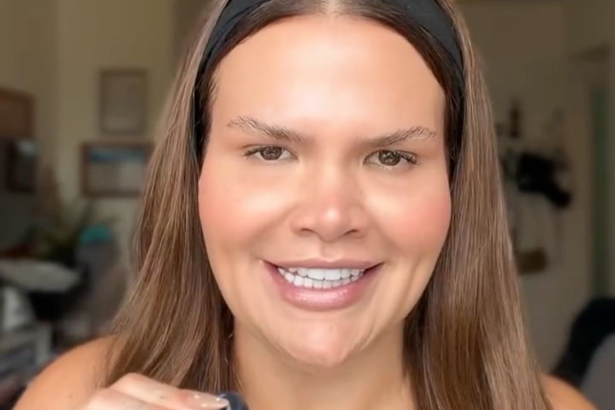 Close de rosto de Thais Carla sorrindo diretamente para a câmera, mostrando dentes brancos e covinhas marcadas. Ela usa uma tiara preta no cabelo castanho solto. A maquiagem é leve e natural. Na parte inferior, vê-se a parte superior de um pequeno frasco e a ponta de um dedo com unha decorada. O fundo é um ambiente interno desfocado.