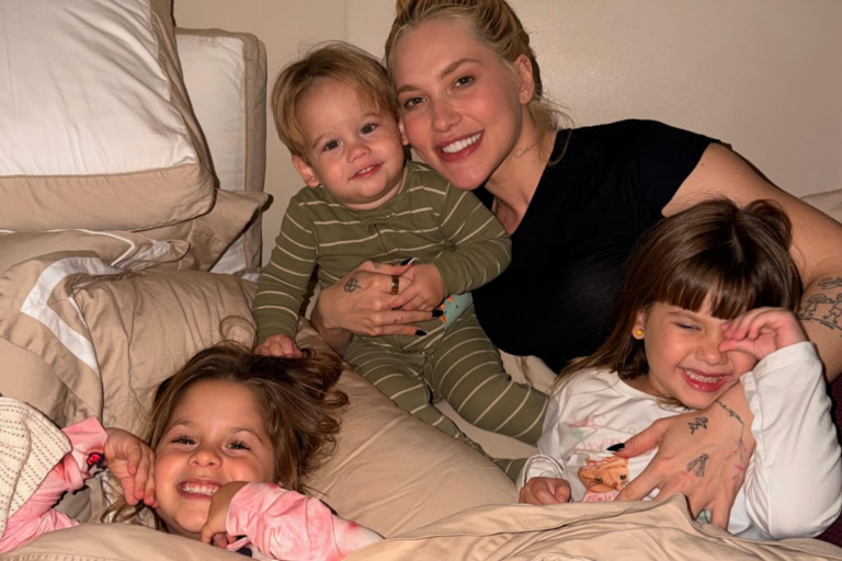 Virginia Fonseca sorridente ao lado de seus três filhos, Maria Alice, Maria Flor e José Leonardo, em um momento descontraído sobre uma cama com lençóis beges.