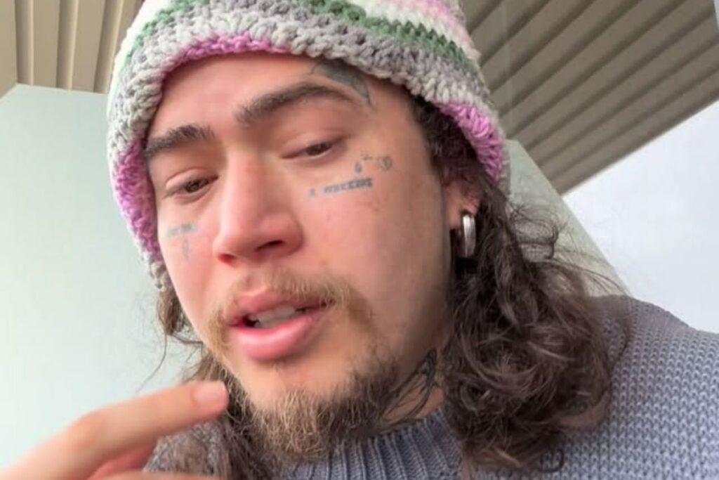 Whindersson Nunes em close-up, com expressão séria, usando uma touca de crochê em tons de cinza, branco e rosa. Ele apresenta barba rala, cabelos longos ondulados e tatuagens faciais visíveis próximas aos olhos. Está vestindo um suéter de tricô cinza e usa um brinco de argola prateada.