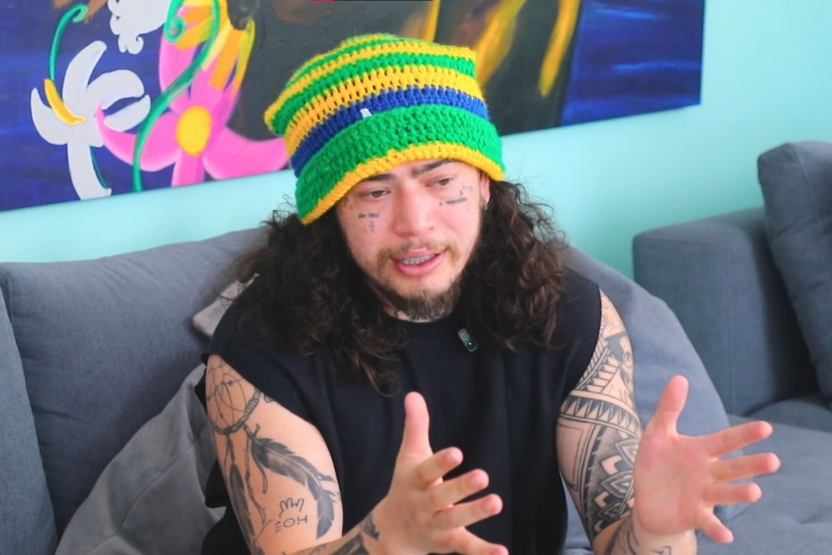 Whindersson Nunes sentado em um sofá cinza, vestindo uma camiseta regata preta e uma touca de crochê com as cores da bandeira do Brasil (verde, amarelo e azul). Ele possui cabelos longos e cacheados, tatuagens visíveis nos braços e no rosto, e aparece gesticulando com as mãos durante uma conversa. Ao fundo, uma tela colorida com detalhes em rosa e azul.