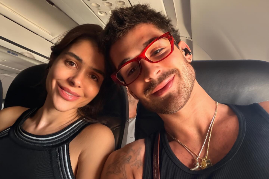 João Guilherme, usando óculos de armação vermelha e regata preta, sorri ao lado de sua mãe, Naira Ávilla, que veste uma blusa preta com detalhes brancos na gola. Ambos estão sentados em poltronas de um avião.