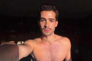 João Guilherme em uma selfie sem camisa, olhando diretamente para a câmera. O ator e influenciador tem uma barba curta e bem aparada, e usa um colar dourado fino com um pequeno pingente retangular. A luz solar bate em seu rosto e torso, enquanto o fundo mostra uma estante de madeira em um ambiente interno.