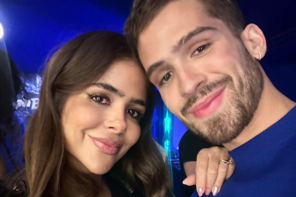 João Guilherme, vestindo uma blusa azul royal, sorri encostado à sua mãe, Naira Ávilla, que possui cabelos castanhos ondulados e usa uma blusa preta. O fundo possui iluminação azulada e é possível ver a mão de Naira com anéis apoiada no ombro dele.