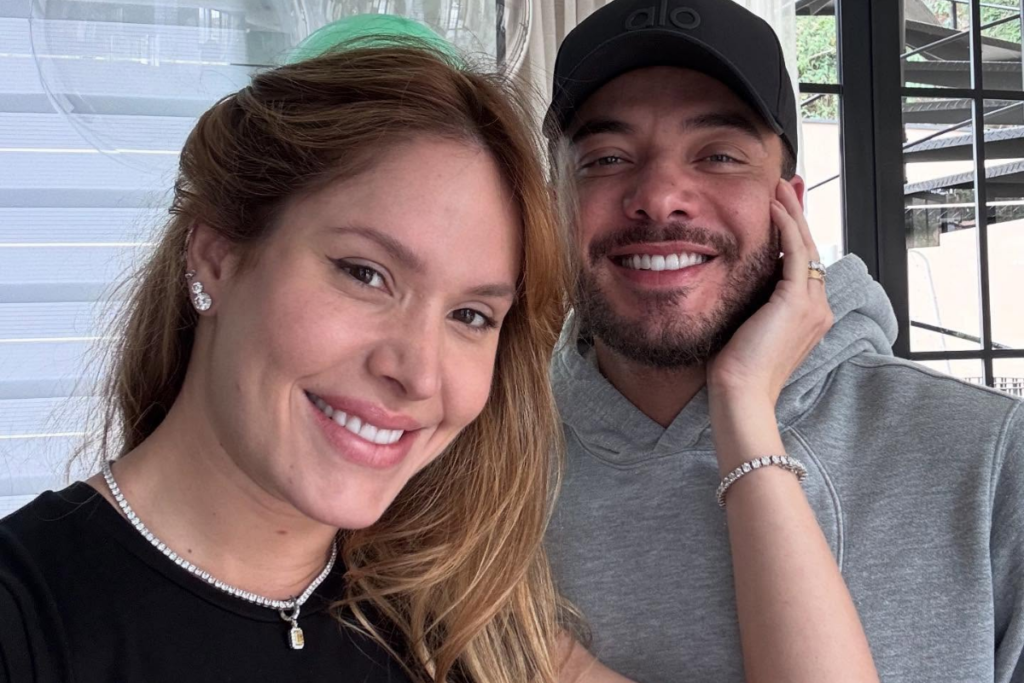 Thyane Dantas sorri para a câmera ao lado de Wesley Safadão. Ela usa uma blusa preta e um colar de brilhantes; ele veste um moletom cinza com capuz e um boné preto, sorrindo enquanto apoia o rosto na mão.
