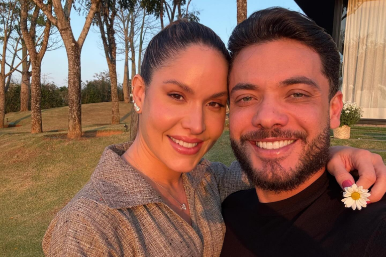 Wesley Safadão e Thyane Dantas sorriem abraçados em um ambiente externo ao entardecer. Thyane, com o cabelo preso em um coque alto e vestindo uma blusa cinza, segura uma pequena flor branca; Wesley usa uma camiseta preta e sorri com o rosto próximo ao dela.