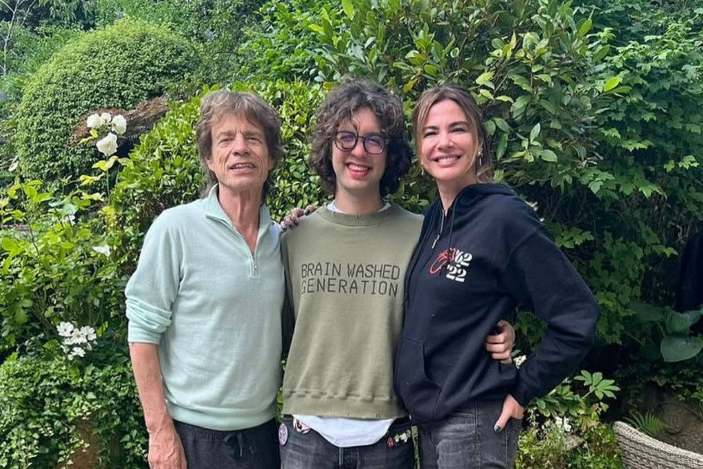 Mick Jagger, Lucas Jagger e Luciana Gimenez posando sorridentes em um jardim frondoso com folhagens verdes ao fundo. Mick Jagger usa uma blusa verde clara, Lucas usa um moletom com a frase
