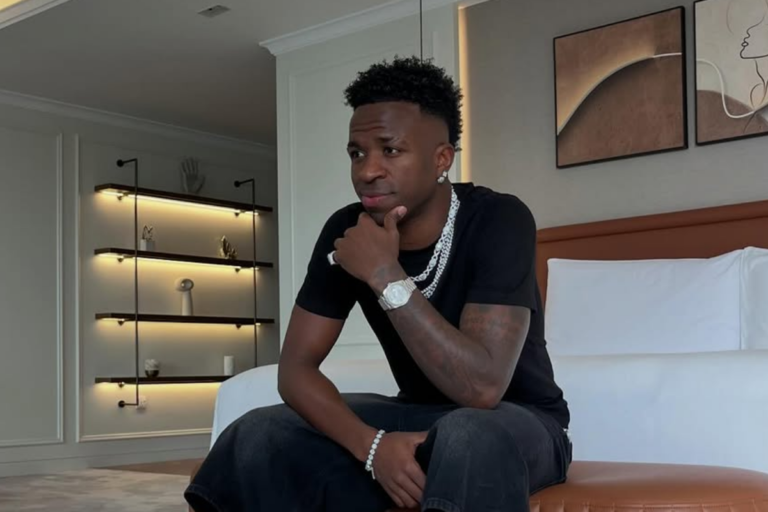 Vinícius Júnior sentado em um banco de couro marrom, vestindo camiseta preta e calças escuras. Ele está com a mão no queixo em pose reflexiva, usando um relógio prateado e colares de corrente. Ao fundo, um quarto moderno com estantes iluminadas e quadros abstratos na parede.