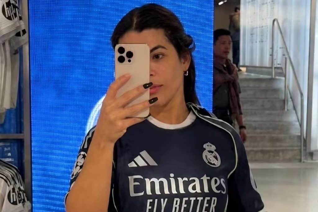 Rosana Ferreira tirando uma selfie em frente a um espelho ou tela azul brilhante. Ela veste uma camisa azul marinho do time Real Madrid com detalhes brancos e patrocínio da Emirates. O celular, de cor clara com três lentes de câmera, cobre parcialmente seu rosto. Suas unhas estão pintadas de preto longo.