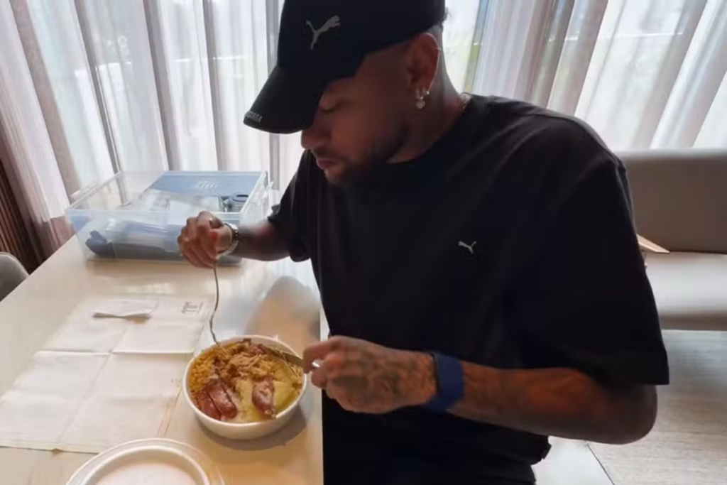 Neymar Jr. usando boné e camiseta preta da Puma, sentado à mesa enquanto come uma marmita composta por arroz, feijão, farofa e carne. Ele segura um garfo e está concentrado na refeição.