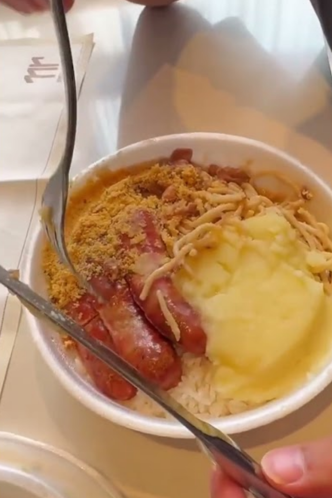Close de uma marmita em prato de esferovite contendo arroz, feijão, puré de batata, farofa e salsichas grelhadas, sendo servida com garfo e faca.