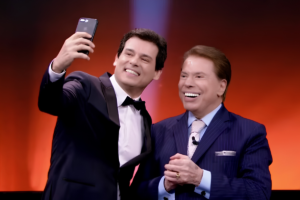 Celso Portiolli sorri ao tirar uma selfie com Silvio Santos no palco; ambos estão de terno e gravata borboleta, com um fundo iluminado em tons de laranja e vermelho.