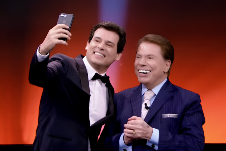 Celso Portiolli sorri ao tirar uma selfie com Silvio Santos no palco; ambos estão de terno e gravata borboleta, com um fundo iluminado em tons de laranja e vermelho.
