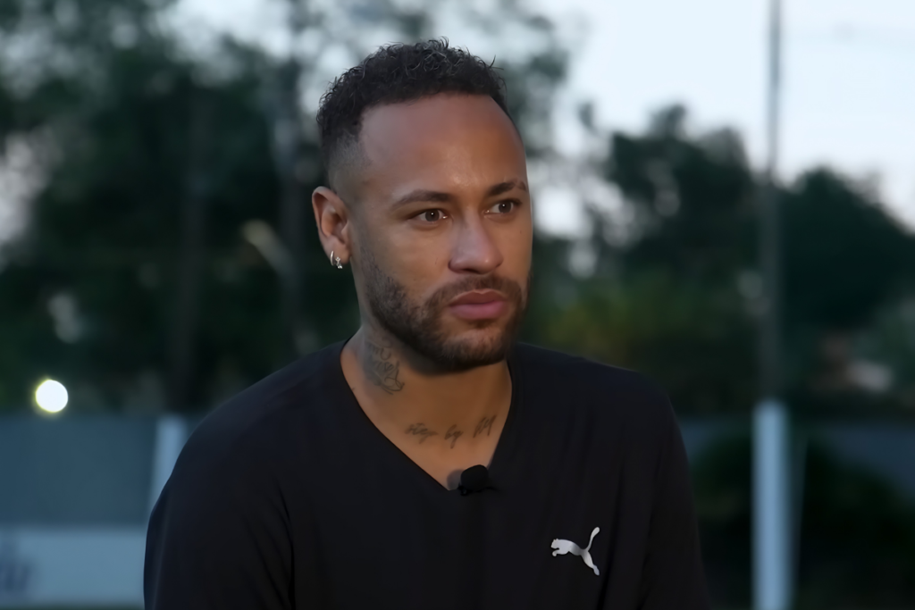 Neymar Jr. de frente, com expressão séria, vestindo uma camiseta preta da Puma com decote em V e microfone de lapela. É possível ver suas tatuagens no pescoço e um brinco pequeno na orelha.