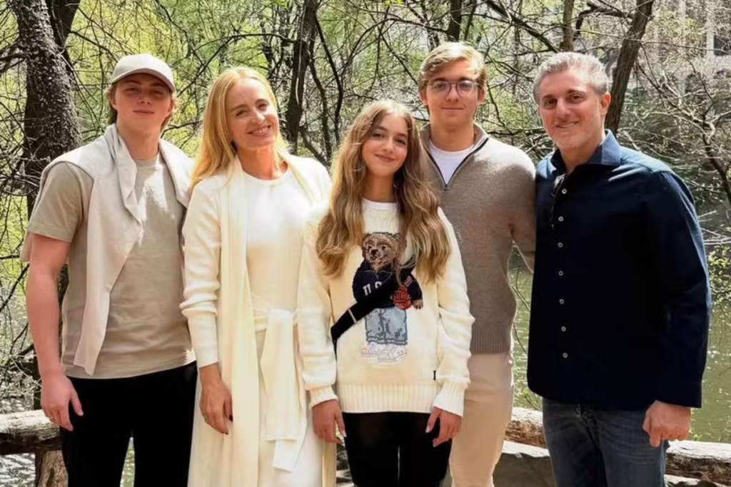 Angélica e Luciano Huck posam sorridentes ao lado dos três filhos, Joaquim, Benício e Eva, em um cenário arborizado ao ar livre.