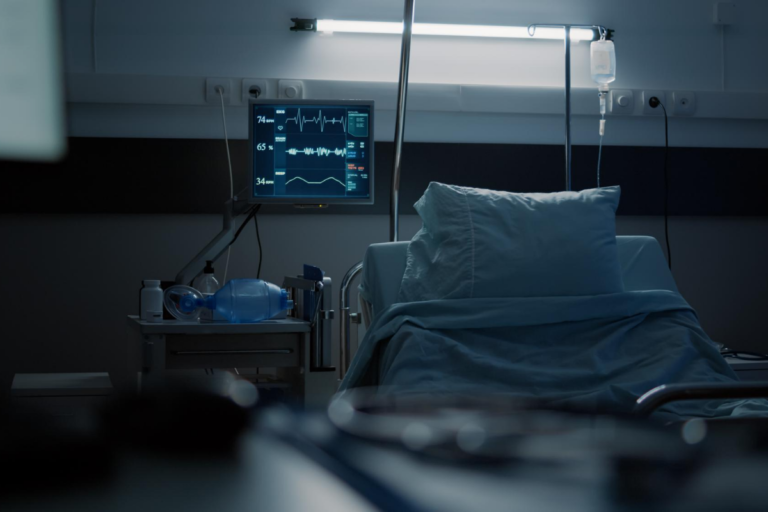 Quarto de hospital escuro e vazio com foco em uma cama médica, monitor de sinais vitais ligado exibindo gráficos cardíacos, suporte de soro e equipamentos de ventilação ao lado.