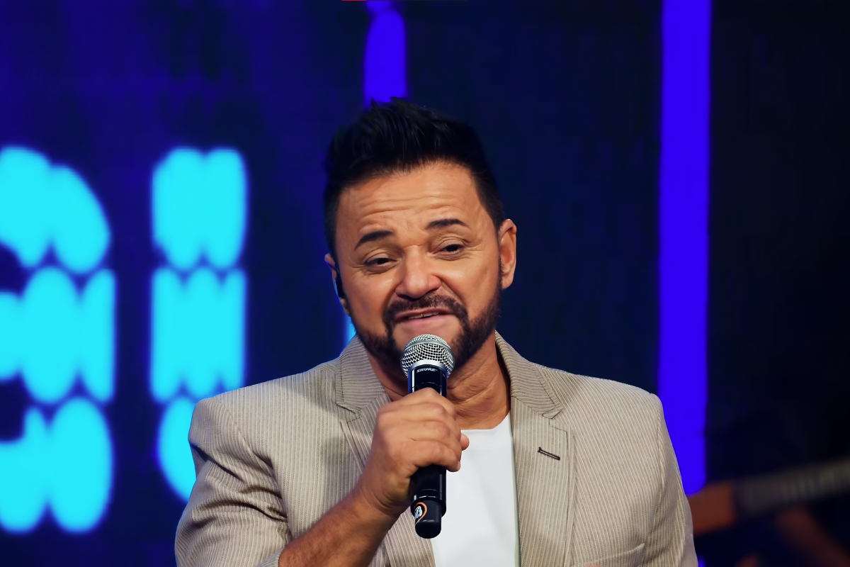 O cantor sertanejo Ray, da dupla Renan e Ray, segurando um microfone próximo à boca durante uma apresentação, vestindo um paletó bege sobre camiseta branca, com luzes de palco azuis ao fundo.