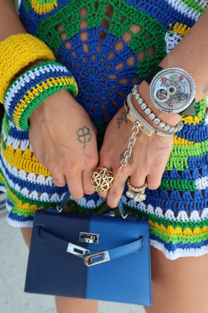 Close nas mãos de Virginia Fonseca exibindo um relógio de luxo Jacob & Co. com diamantes, pulseiras e anéis, vestindo um conjunto de crochê com as cores do Brasil e segurando uma bolsa mini azul.