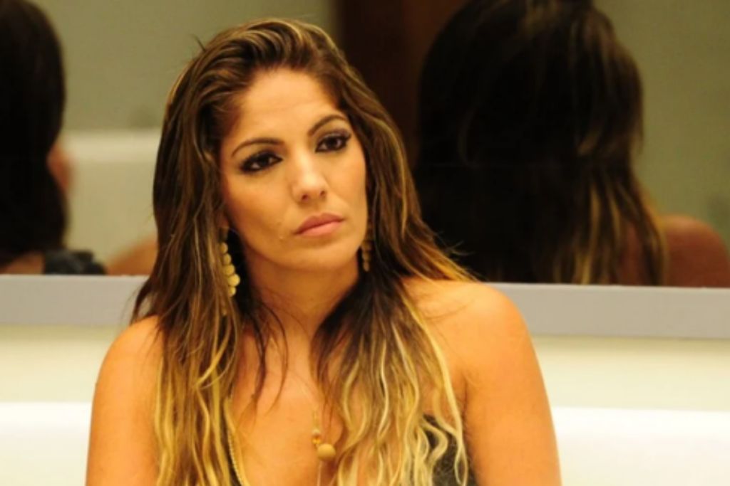 A ex-BBB Anamara aparece em plano médio com olhar atento, usando cabelos longos com mechas claras e brincos pendentes, em um ambiente interno com espelho ao fundo.