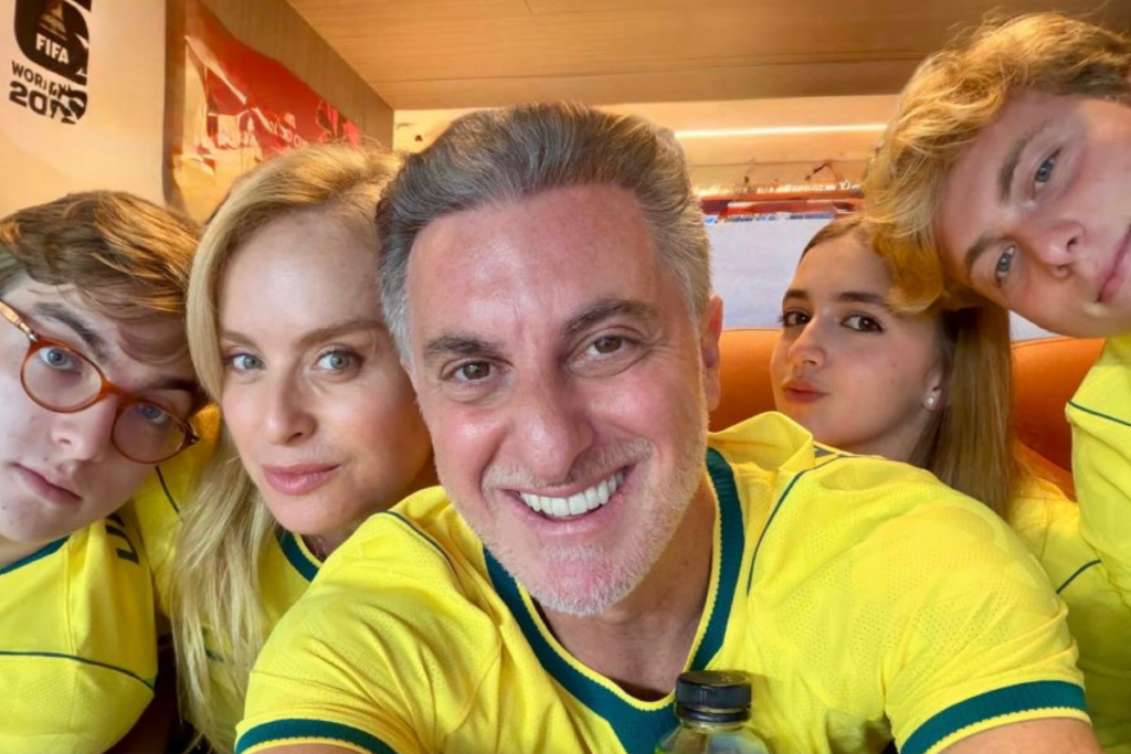 Angélica, Luciano Huck e seus três filhos, Joaquim, Benício e Eva, sorridentes e reunidos para uma selfie. Todos vestem camisas da Seleção Brasileira de Futebol. Ao fundo, detalhes de um evento ou local de visualização da Copa do Mundo de 2022.