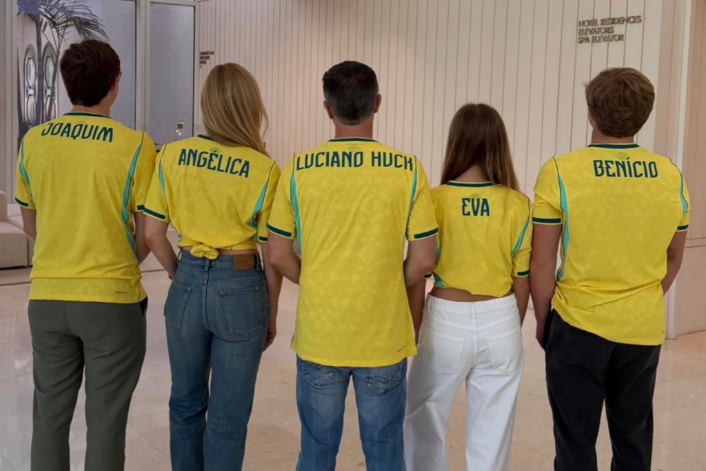 Joaquim, Angélica, Luciano Huck, Eva e Benício de costas, usando camisas da Seleção Brasileira com seus nomes estampados nas costas, em ambiente interno.