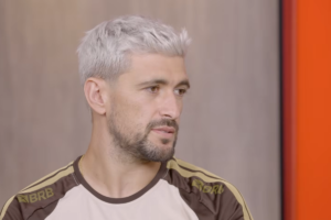 Jogador de futebol Arrascaeta de perfil, com cabelo curto descolorido em tom platinado e barba aparada, vestindo uma camisa esportiva com detalhes em marrom e dourado com a marca BRB, contra um fundo neutro cinza e uma faixa vertical laranja à direita.