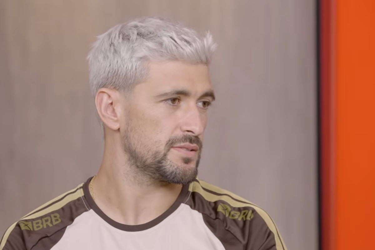 Jogador de futebol Arrascaeta de perfil, com cabelo curto descolorido em tom platinado e barba aparada, vestindo uma camisa esportiva com detalhes em marrom e dourado com a marca BRB, contra um fundo neutro cinza e uma faixa vertical laranja à direita.