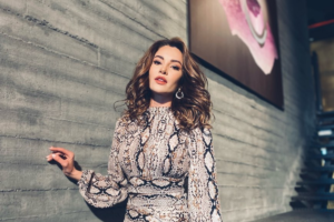 Atriz Ece İrtem posa encostada em parede de concreto com look de estampa animal print, usando cabelos ondulados e brincos circulares.
