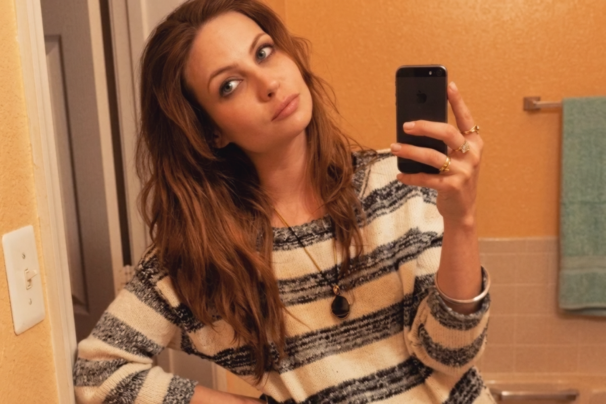Atriz Daveigh Chase posando para uma selfie em frente ao espelho usando um suéter listrado e um colar com pingente escuro.