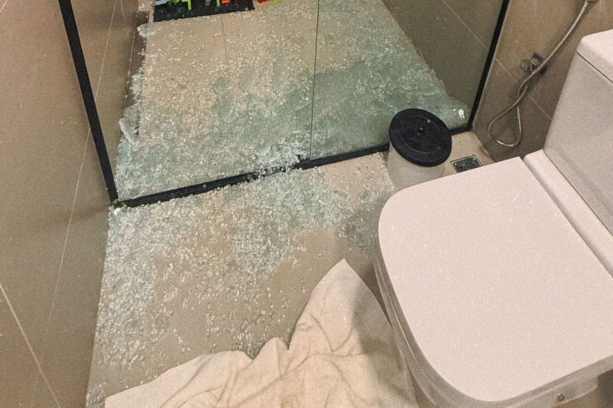 Chão de banheiro coberto por milhares de estilhaços de vidro temperado verde após o box estourar no acidente do ex-BBB Breno Simões. Ao lado, aparecem parte do vaso sanitário branco e uma toalha no piso.