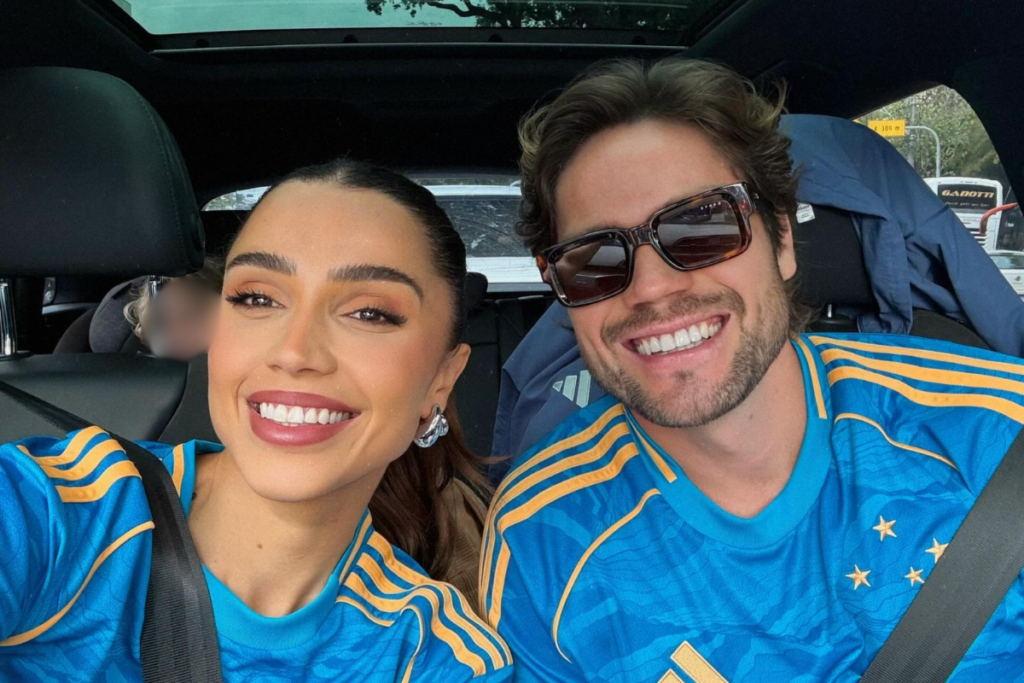 Os ex-BBBs Paula Amorim e Breno Simões sorrindo lado a lado dentro de um carro. Ambos vestem camisas de futebol azuis com listras amarelas nos ombros, do time Cruzeiro. Paula está à esquerda com o cabelo preso, brincos prateados e batom marcante, enquanto Breno está à direita usando óculos de sol escuros de armação tartaruga.