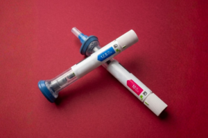 Duas canetas de aplicação de medicamento injetável para perda de peso cruzadas sobre uma superfície de fundo vermelho escuro. A caneta superior possui detalhes em azul com a inscrição 12.5 mg, e a caneta inferior apresenta detalhes em rosa com a inscrição 10 mg.