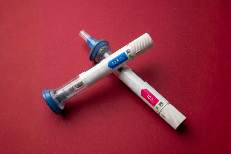 Duas canetas de aplicação de medicamento injetável para perda de peso cruzadas sobre uma superfície de fundo vermelho escuro. A caneta superior possui detalhes em azul com a inscrição 12.5 mg, e a caneta inferior apresenta detalhes em rosa com a inscrição 10 mg.