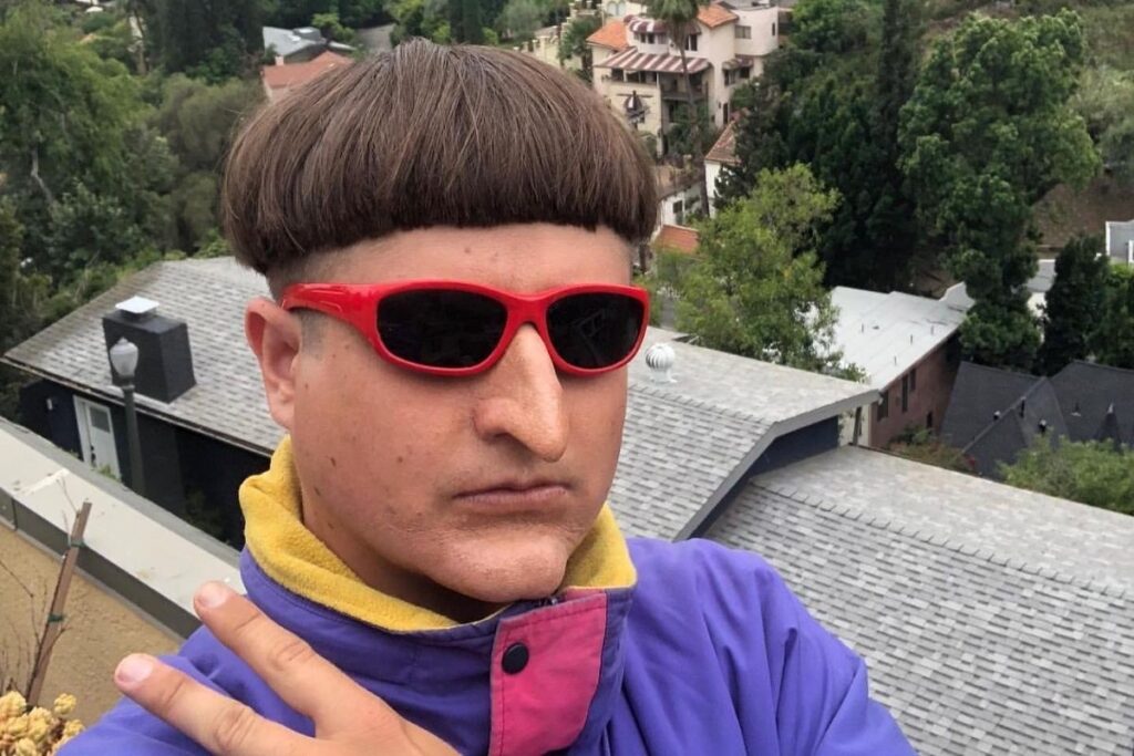Oliver Tree com seu característico corte de cabelo tigela e óculos escuros vermelhos, fazendo um sinal de paz enquanto veste uma jaqueta roxa, com casas e colinas de Los Angeles ao fundo.