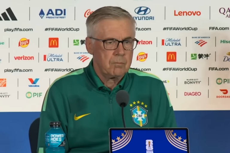 Carlo Ancelotti de óculos, vestindo jaqueta verde da Seleção Brasileira com o escudo da CBF, sentado em frente a um microfone e painel com patrocinadores da Copa do Mundo 2026.