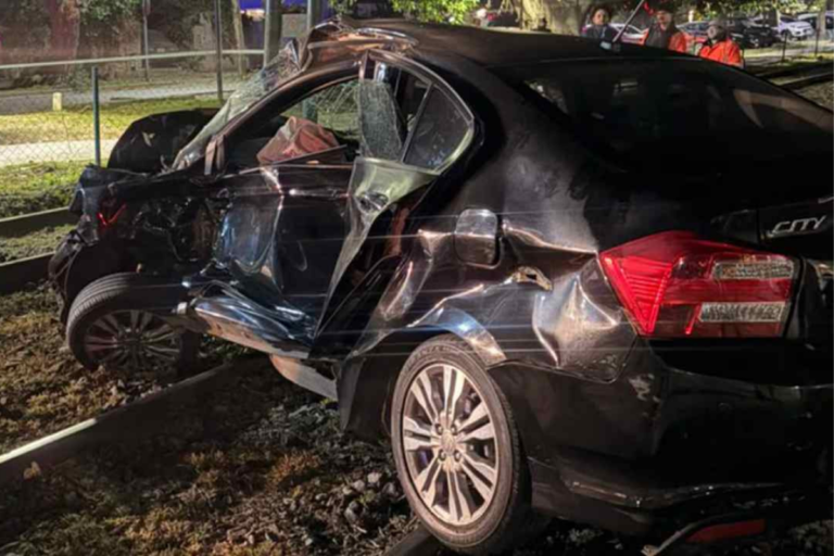 Carro sedan preto severamente danificado após colisão, posicionado sobre trilhos de trem em área externa à noite.