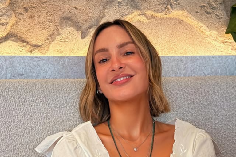 Cantora Claudia Leitte sorri de frente para a câmera. Ela tem cabelos loiros curtos no corte bob, usa blusa branca com decote e colares delicados.