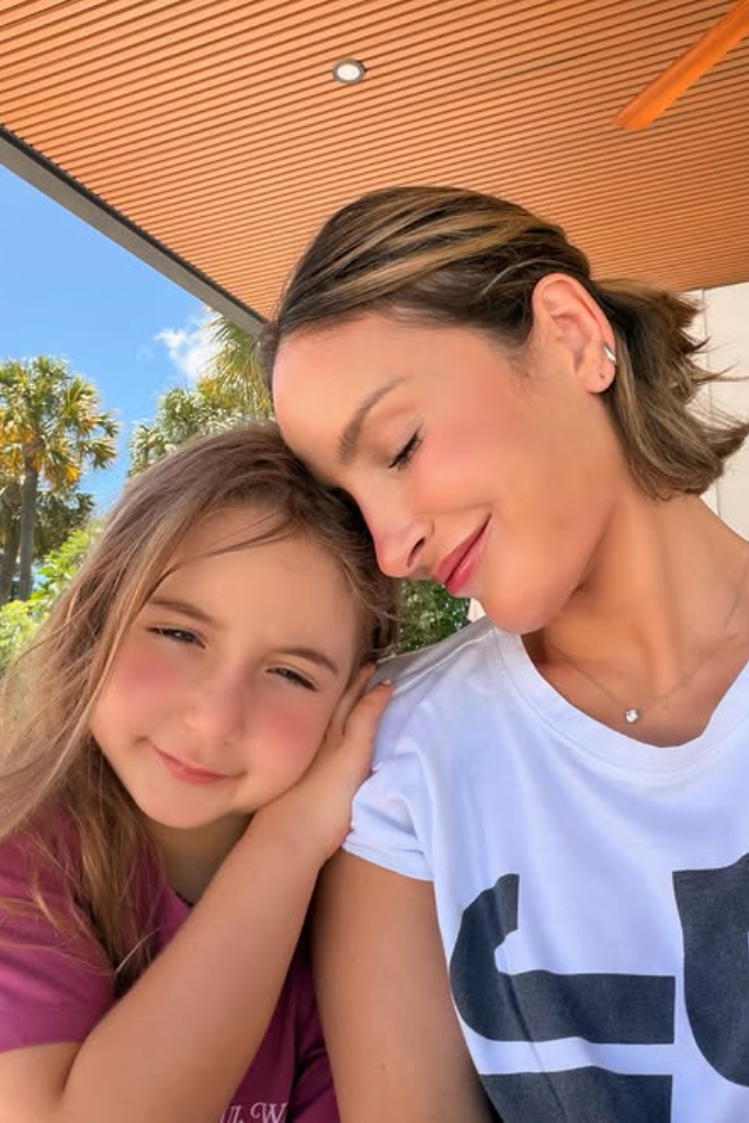 Cantora Claudia Leitte sorri de olhos fechados encostando sua cabeça delicadamente na cabeça de sua filha, Bela, que sorri para a foto apoiando a mão no ombro da mãe.