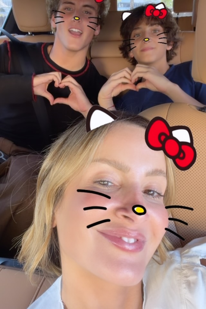 Claudia Leitte sorri em primeiro plano dentro de um carro, enquanto seus dois filhos, Davi e Rafael, aparecem no banco de trás fazendo corações com as mãos. Todos usam um filtro digital de gatinho.